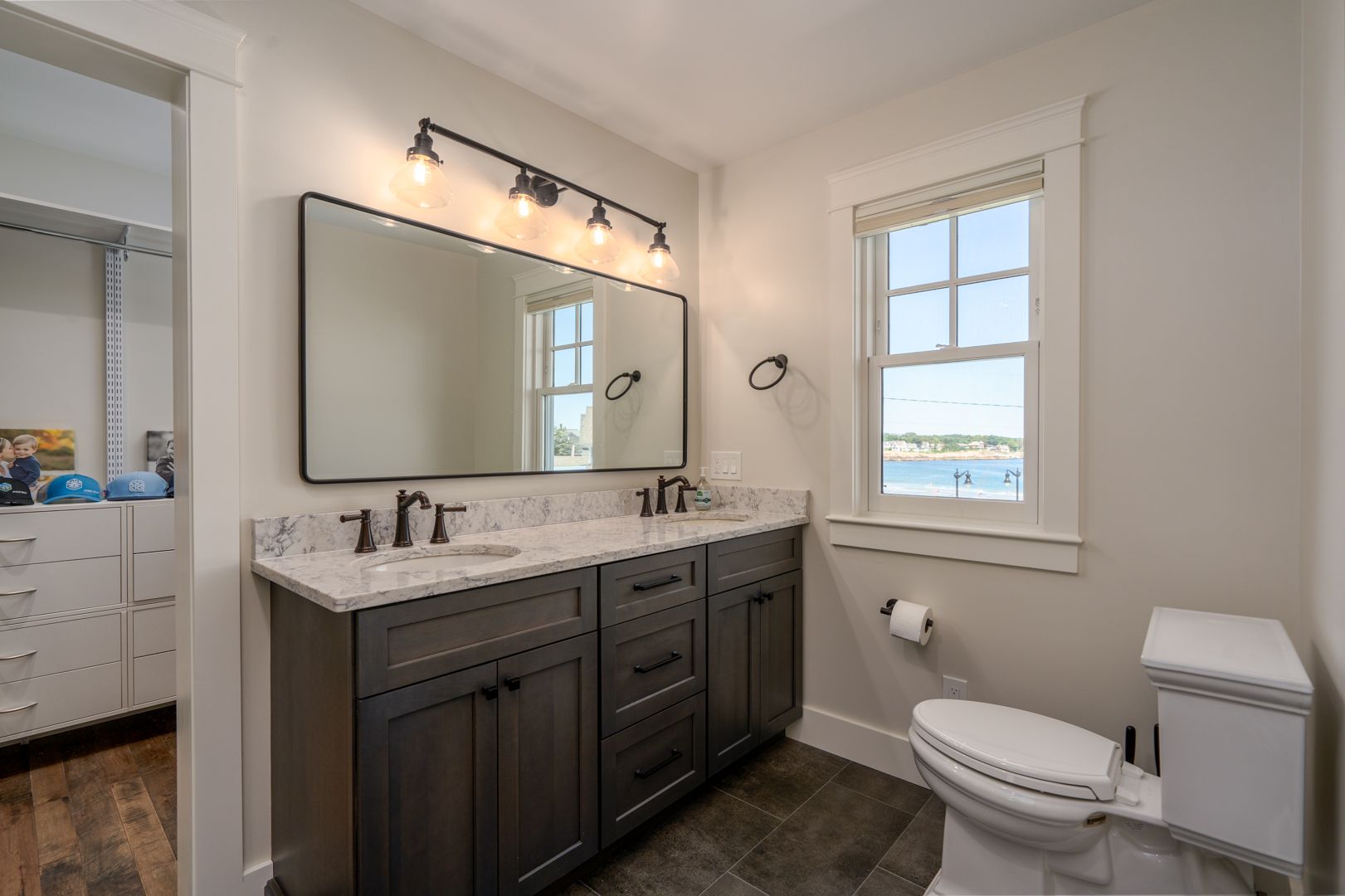 Modern bathroom with dark wood vanity, large mirror, toilet, and window overlooking water.