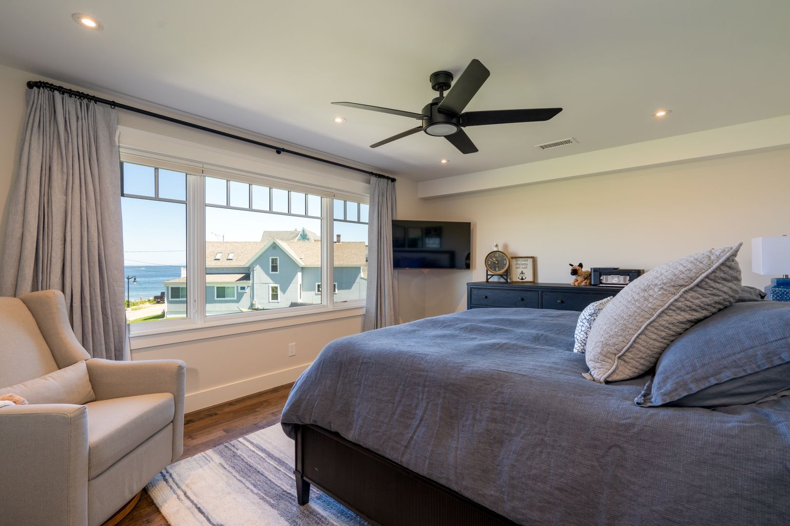 Bright bedroom with gray bed, beige chair, large window, and ceiling fan overlooking the water