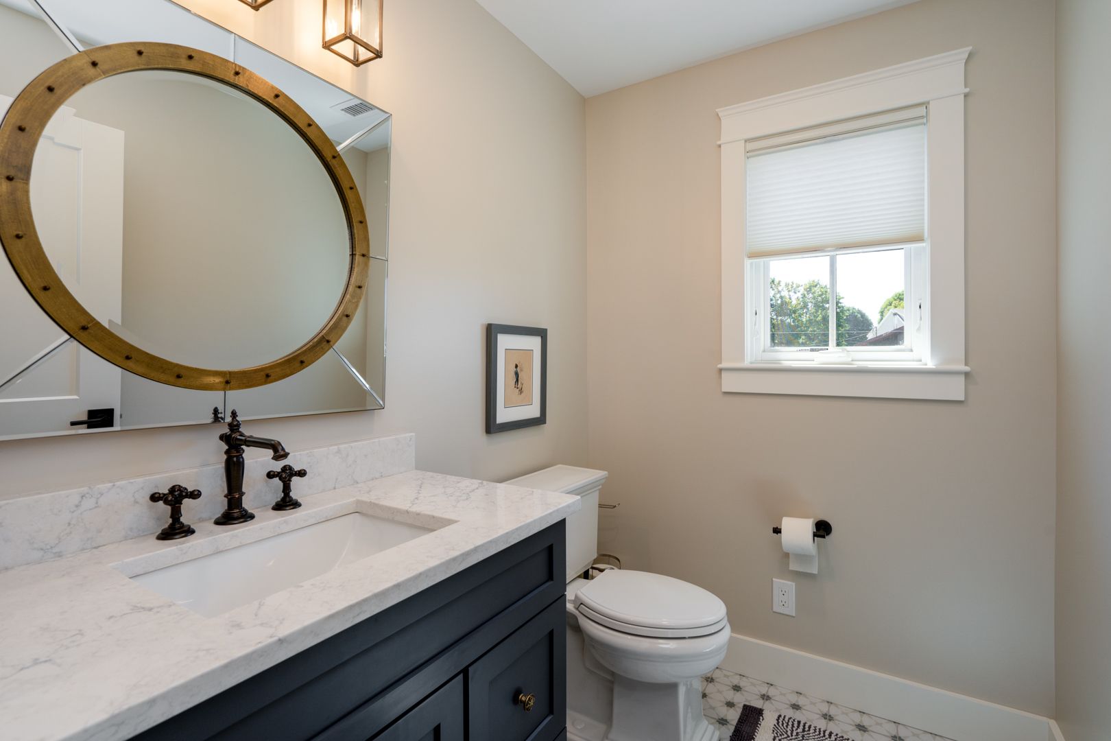 Small bathroom with dark vanity, round mirror, window, and toilet beside marble countertop.