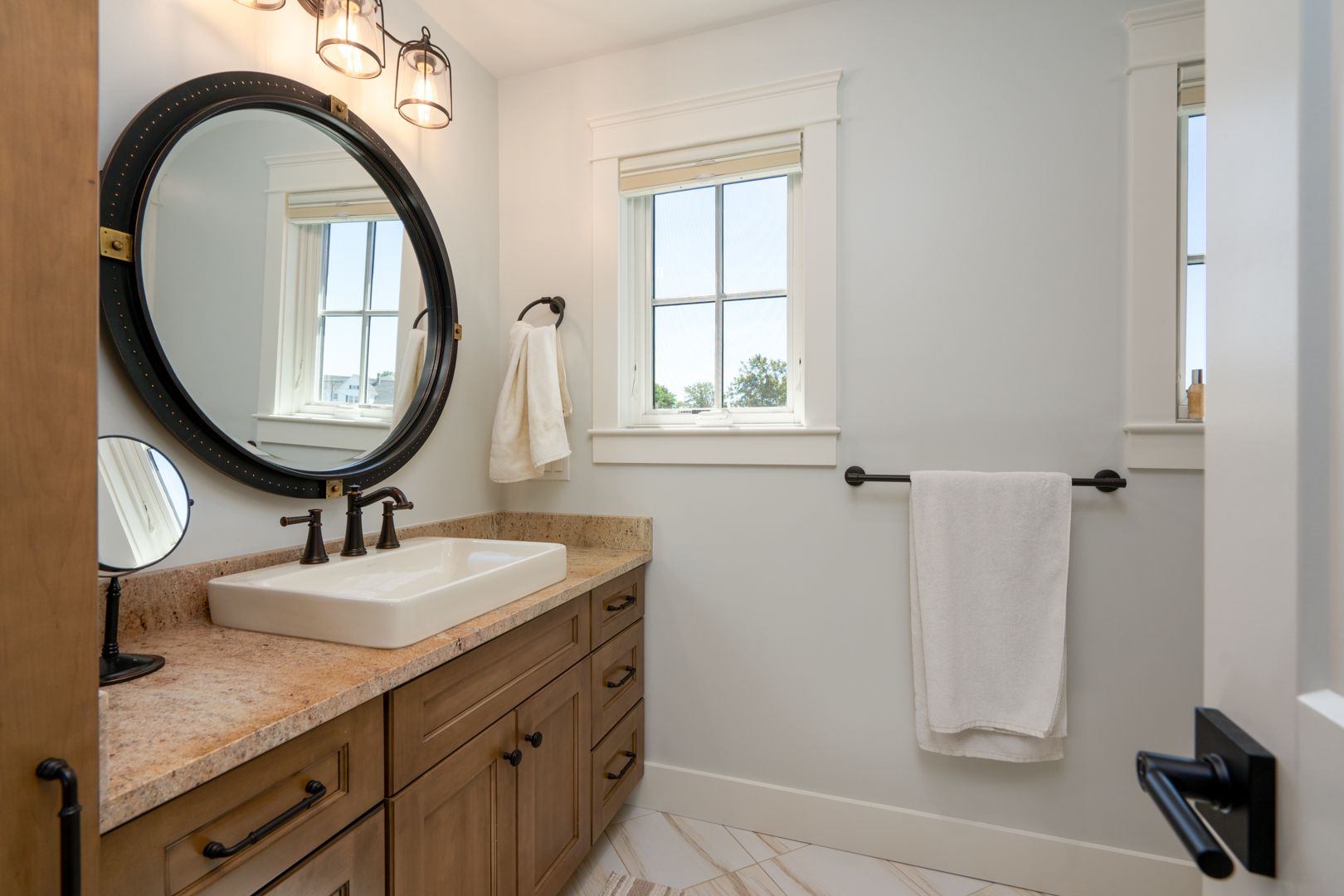 Bright bathroom with round mirror, wood vanity, white sink, and towels by a window