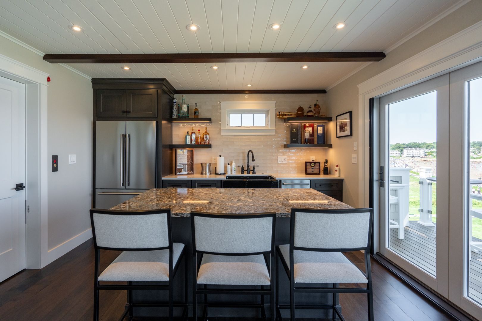 Modern kitchen with granite island, three chairs, stainless fridge, open shelves, and sliding glass doors