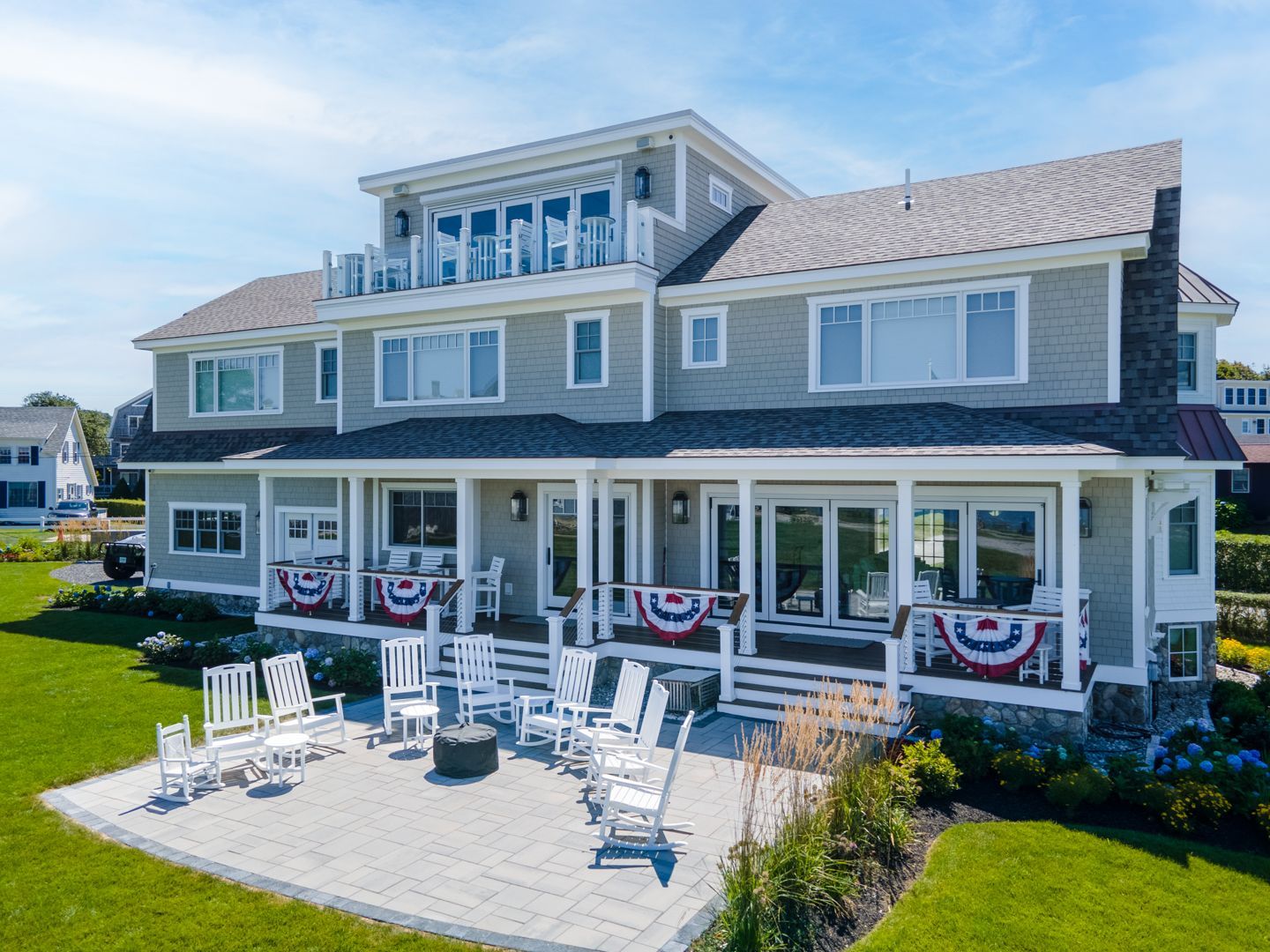 Large gray beachfront house with a white deck, patio chairs, and patriotic bunting on the porch