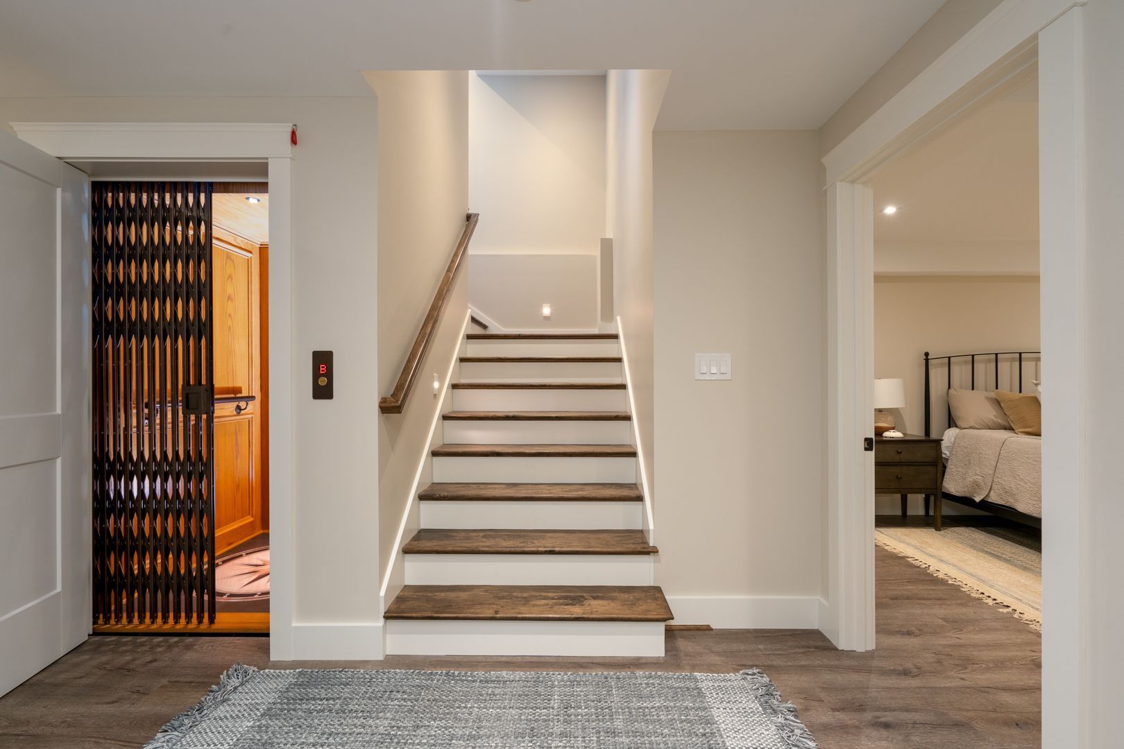 Bright hallway with a central staircase, open elevator on the left, and bedroom visible on the right.