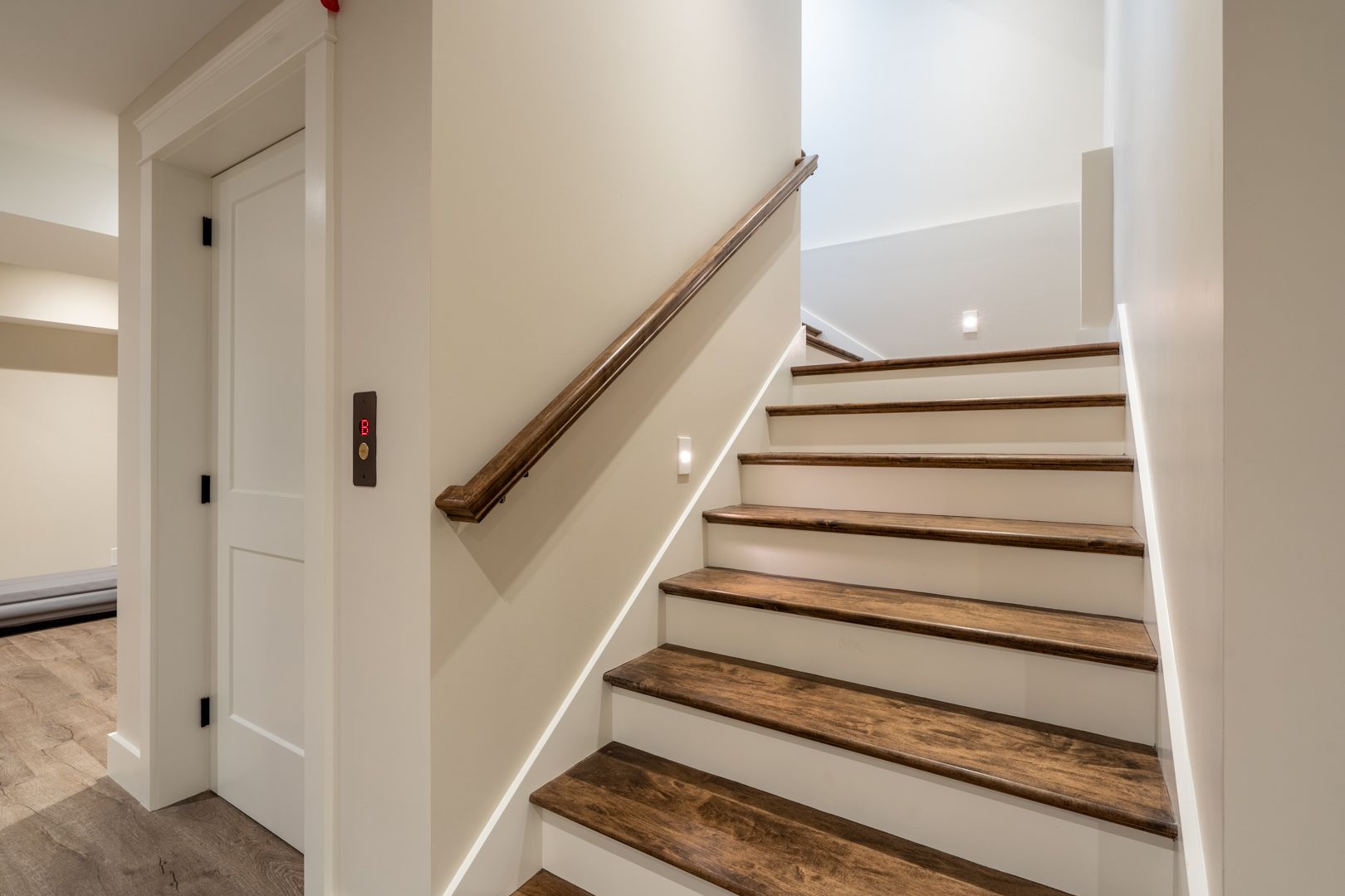Indoor staircase with wood steps and beige walls, viewed from a hallway with a white door and handrail