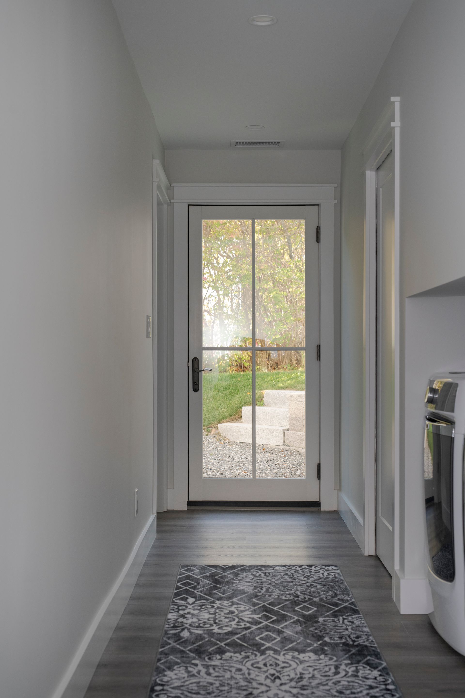 Narrow white hallway with patterned rug leading to a glass door and sunlit backyard beyond.