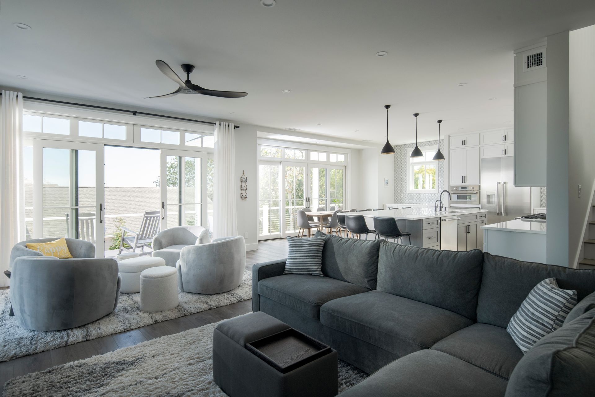 Bright modern living room with gray sectional, armchairs, large windows, and open kitchen.