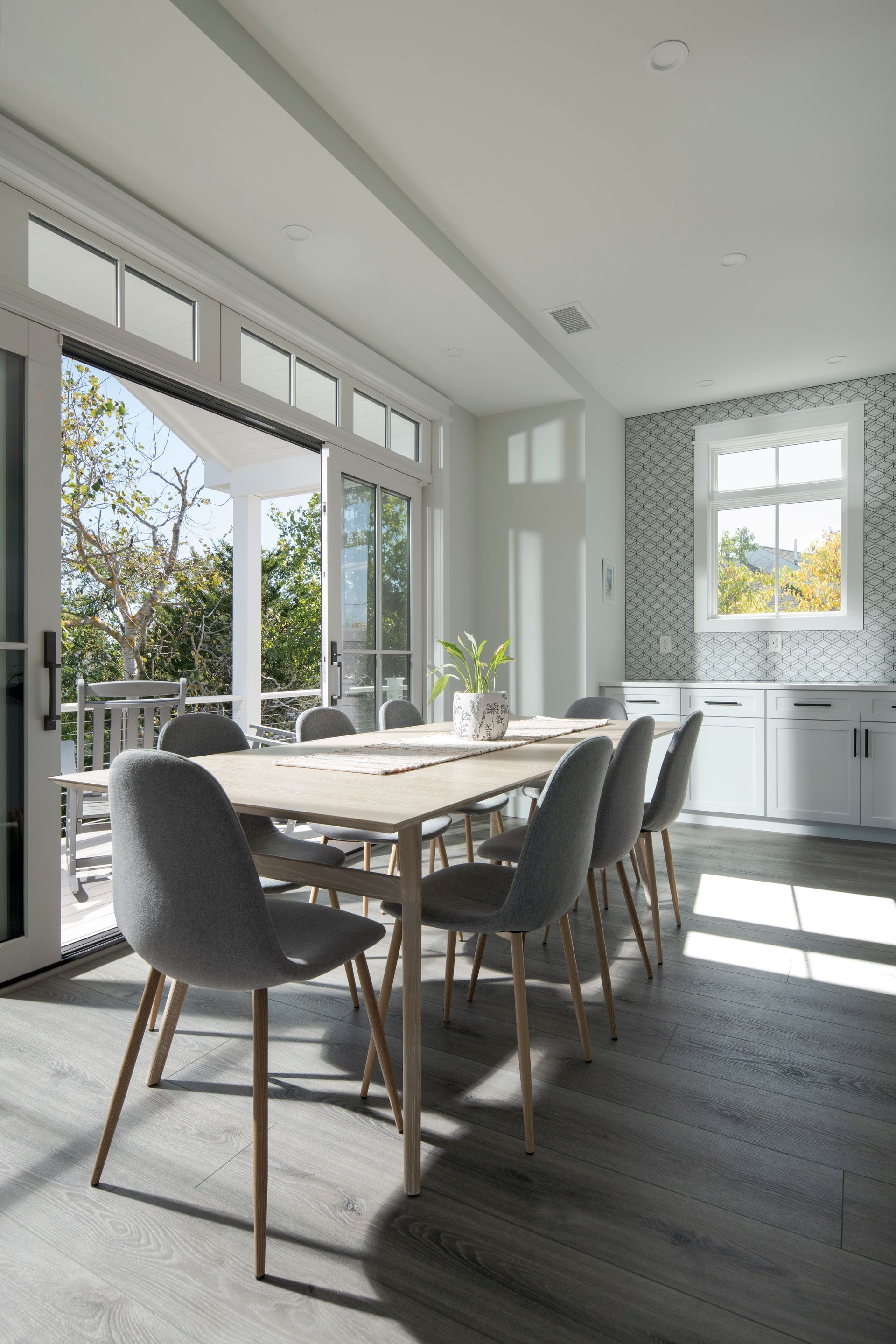 Bright dining room with a wooden table, gray chairs, large windows, and sunlight on dark floors.