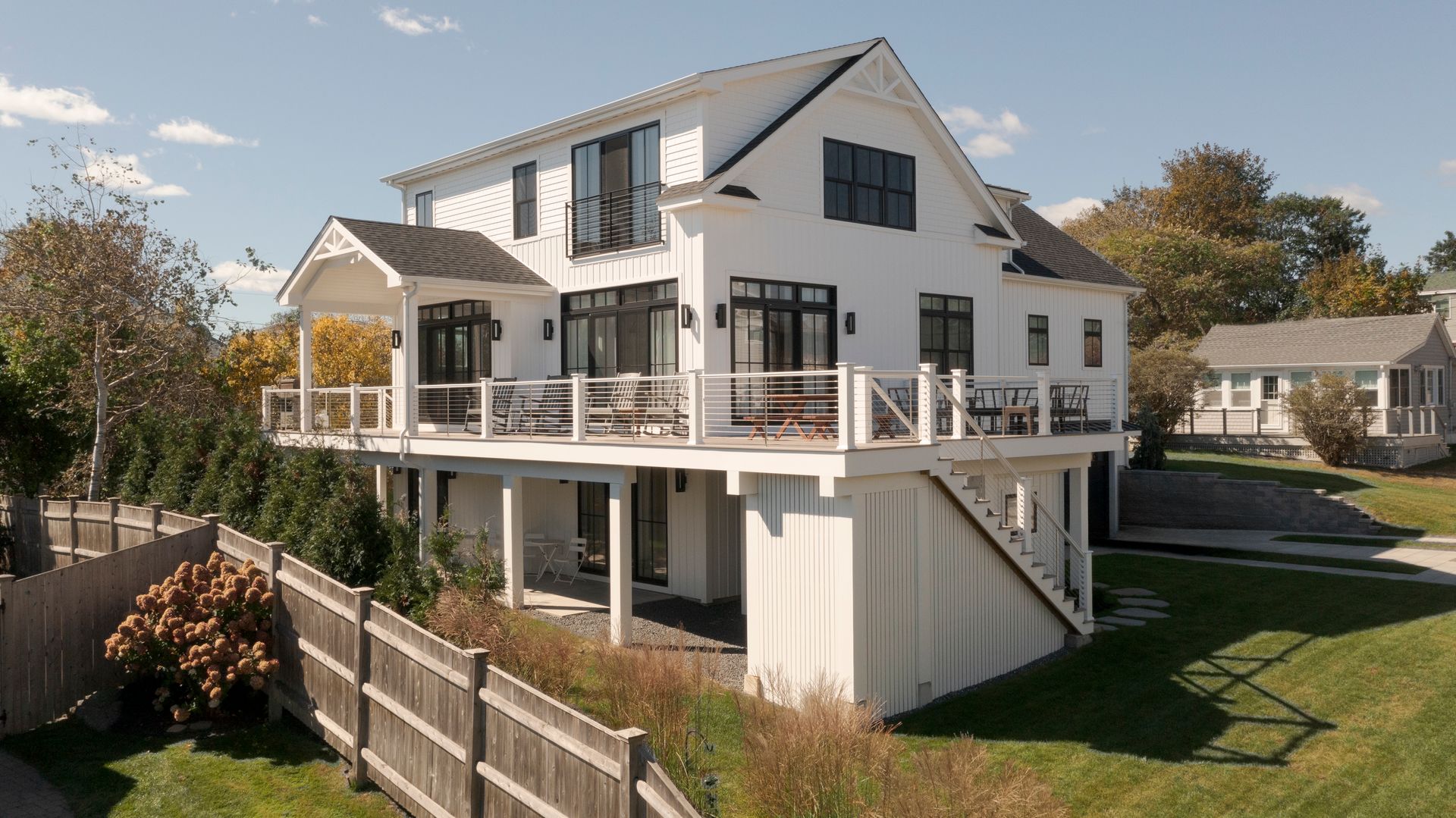 White two-story house with wraparound deck, staircase, and fenced backyard under a blue sky