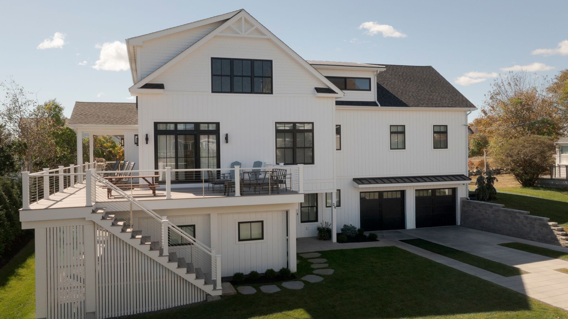 Modern white two-story house with black trim, raised deck, and garage doors on a sunny lawn