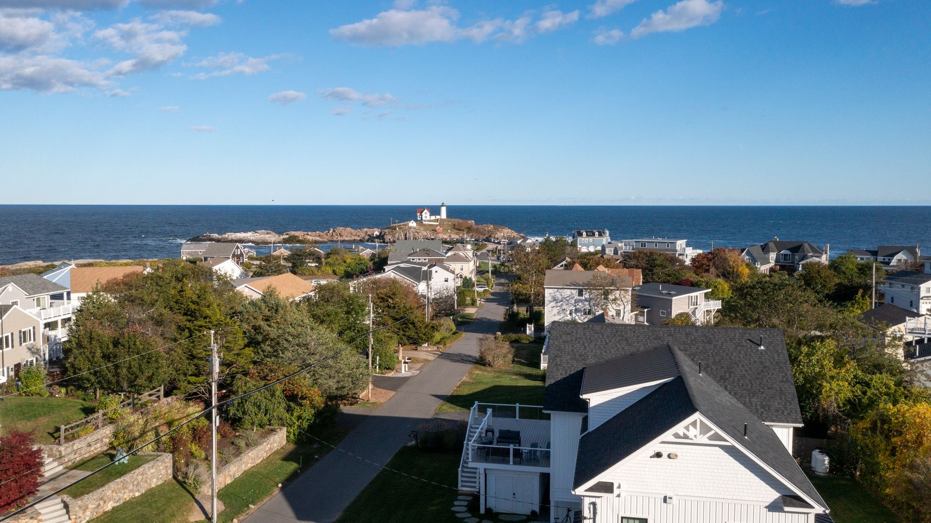 Coastal neighborhood with houses, trees, and a road overlooking the ocean under a partly cloudy sky