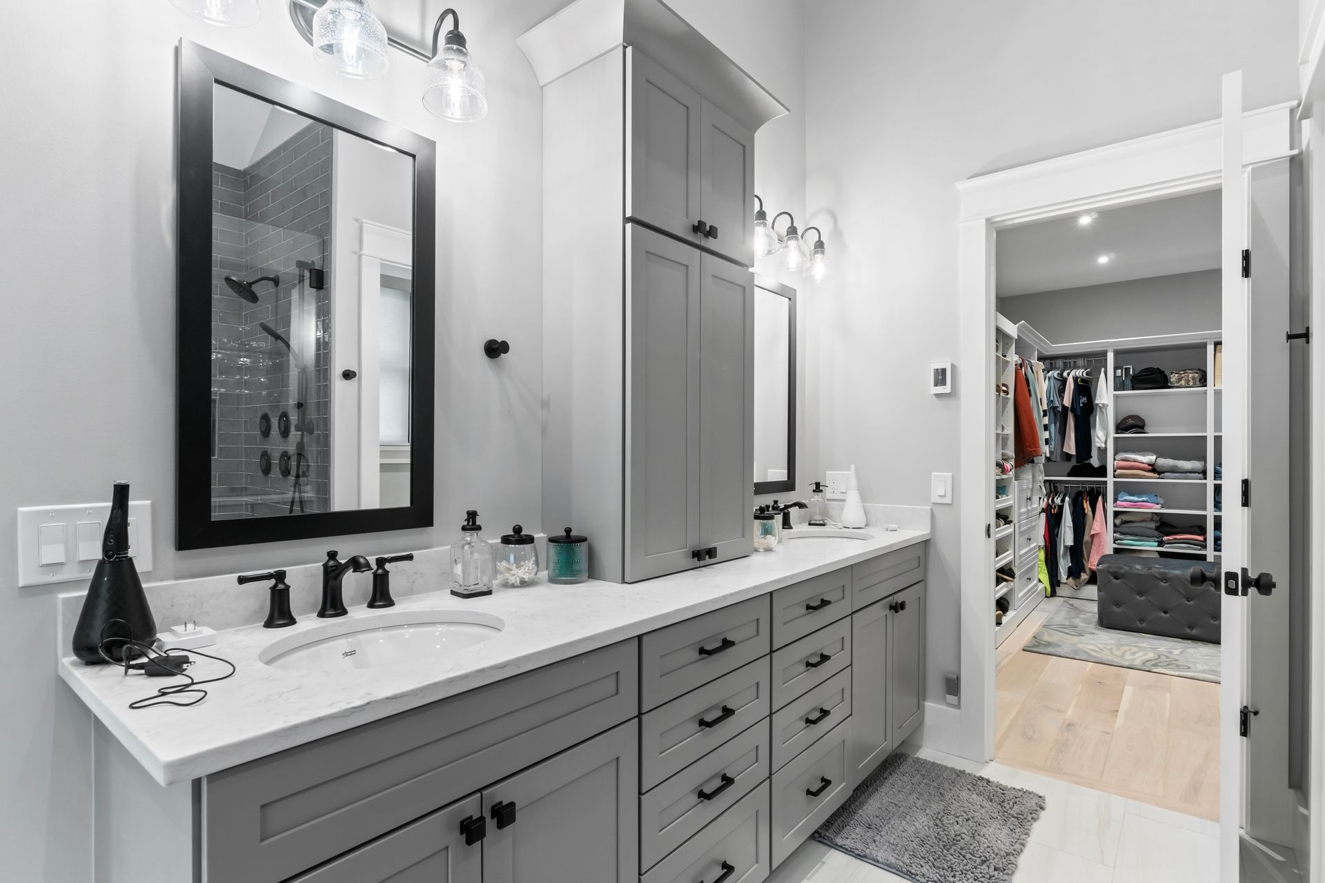Modern bathroom vanity with double sinks, gray cabinets, mirror, and open walk-in closet beyond the doorway