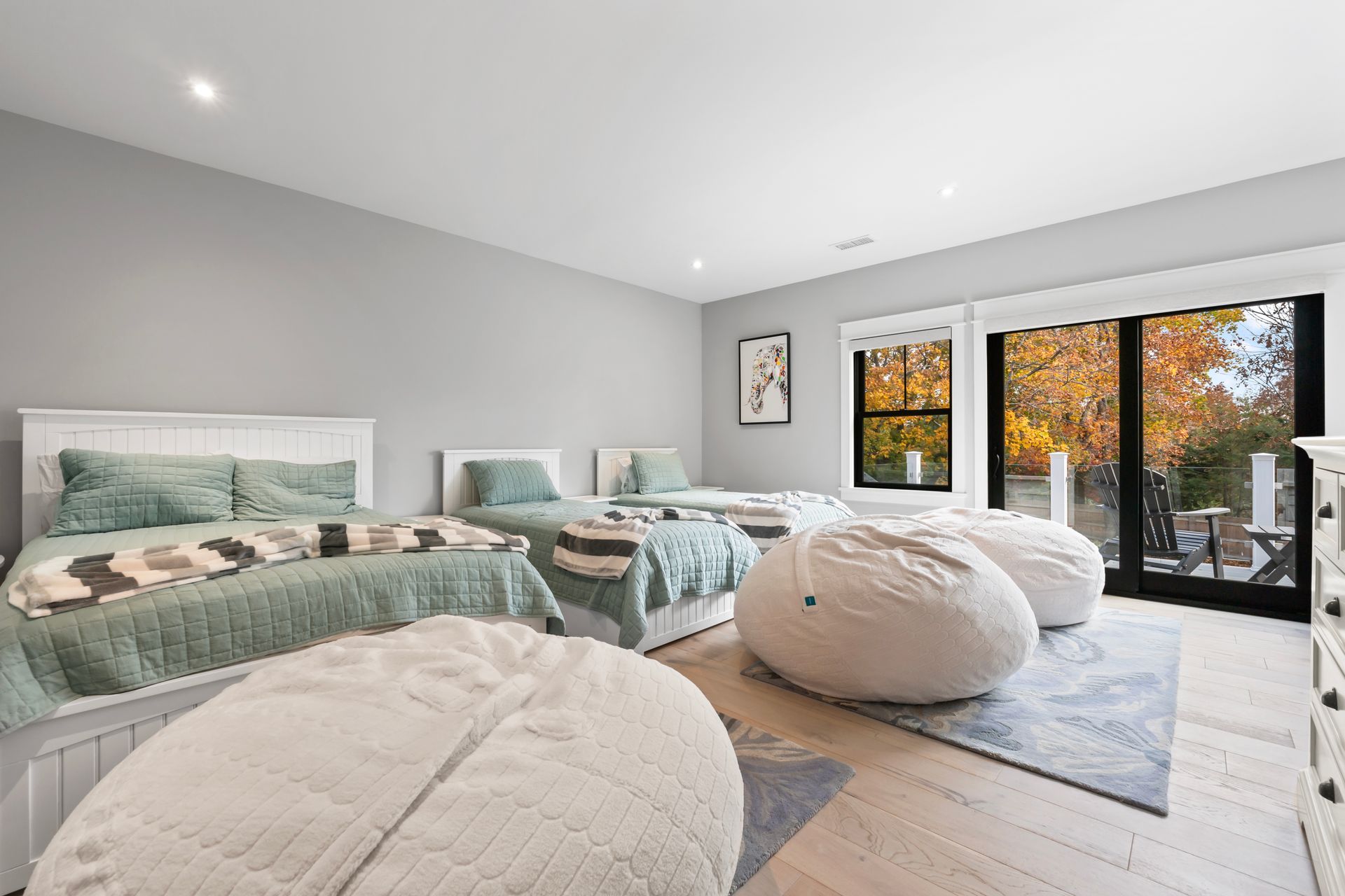 Bright bedroom with three beds, beanbag chairs, and large windows showing autumn trees.