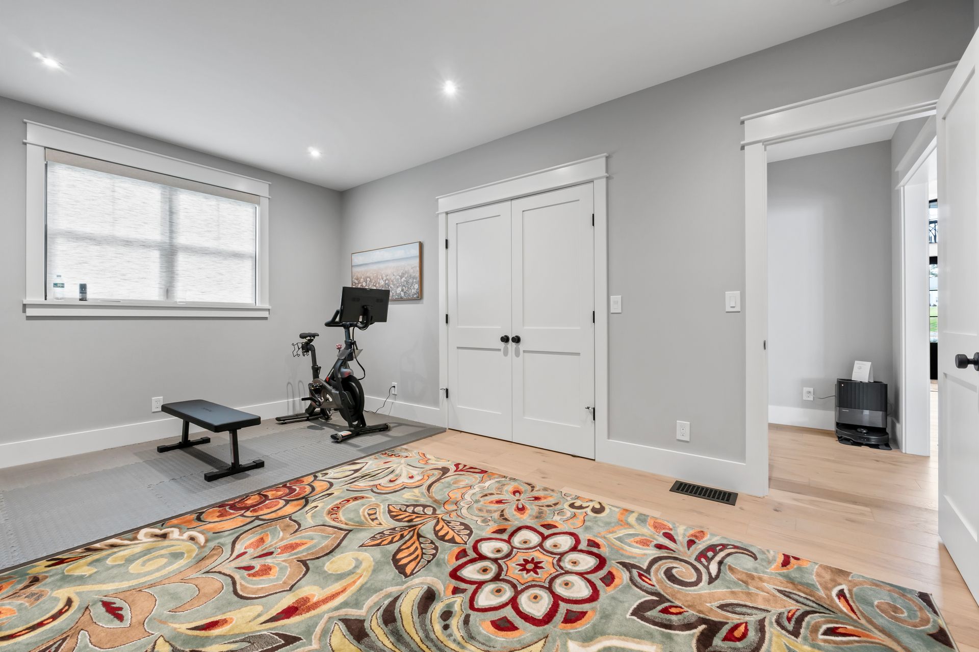 Bright home gym with bench, stationary bike, patterned rug, and double closet doors in a gray room