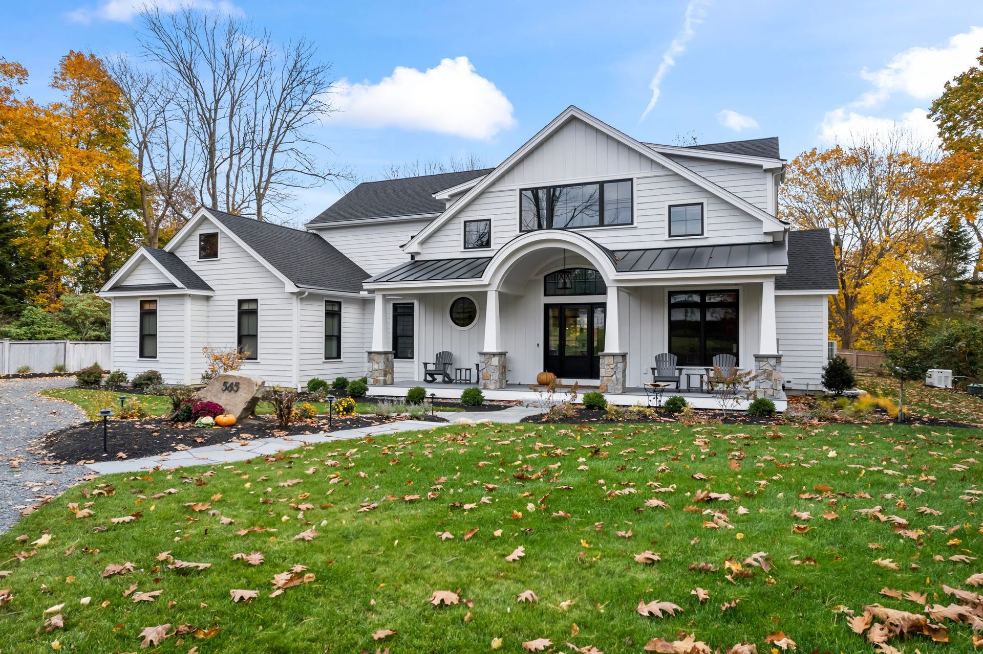 White two-story suburban house with arched porch, landscaped yard, and autumn trees under a blue sky