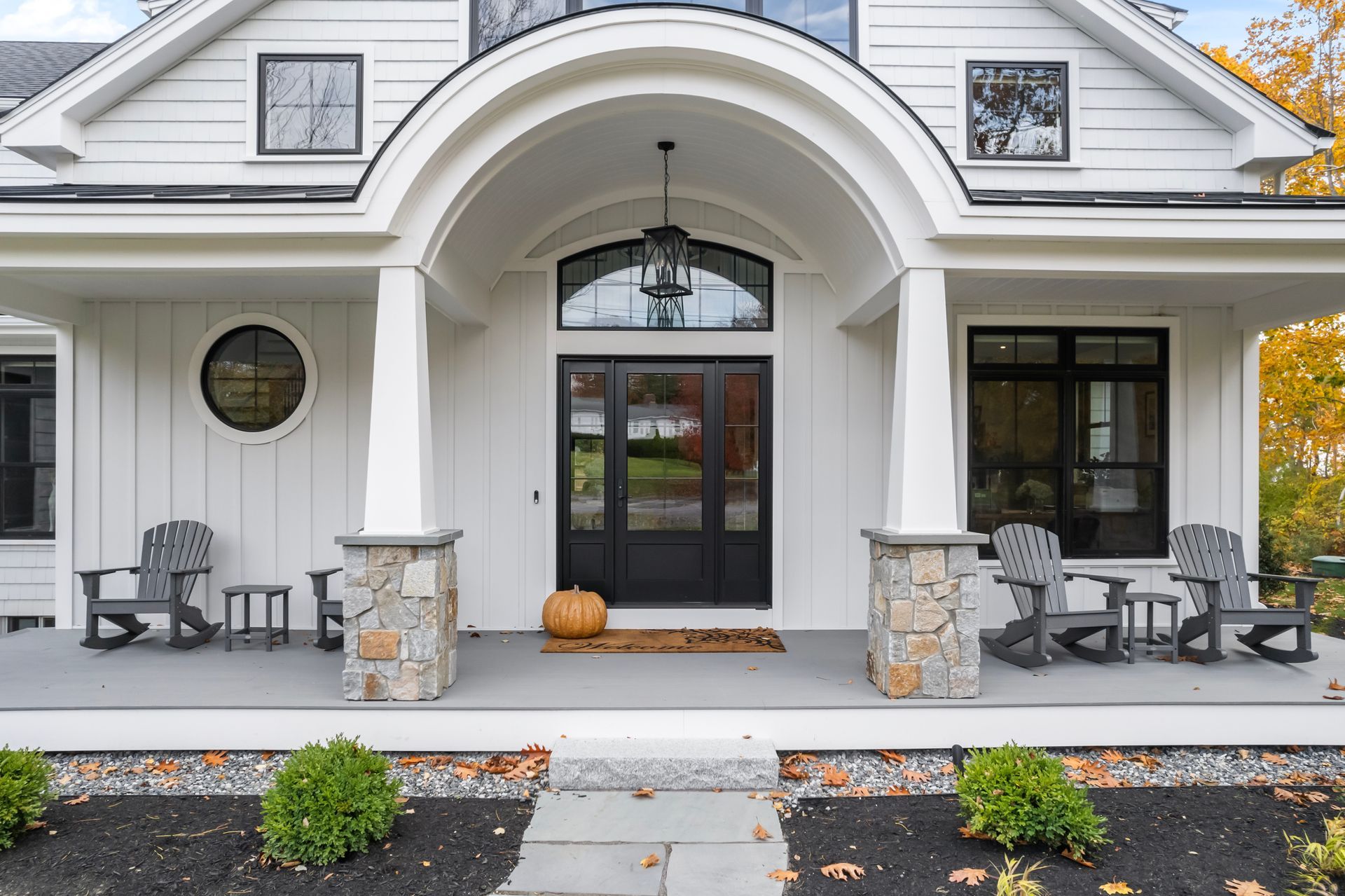 Front porch of a white house with stone pillars, black double doors, chairs, and fall pumpkins