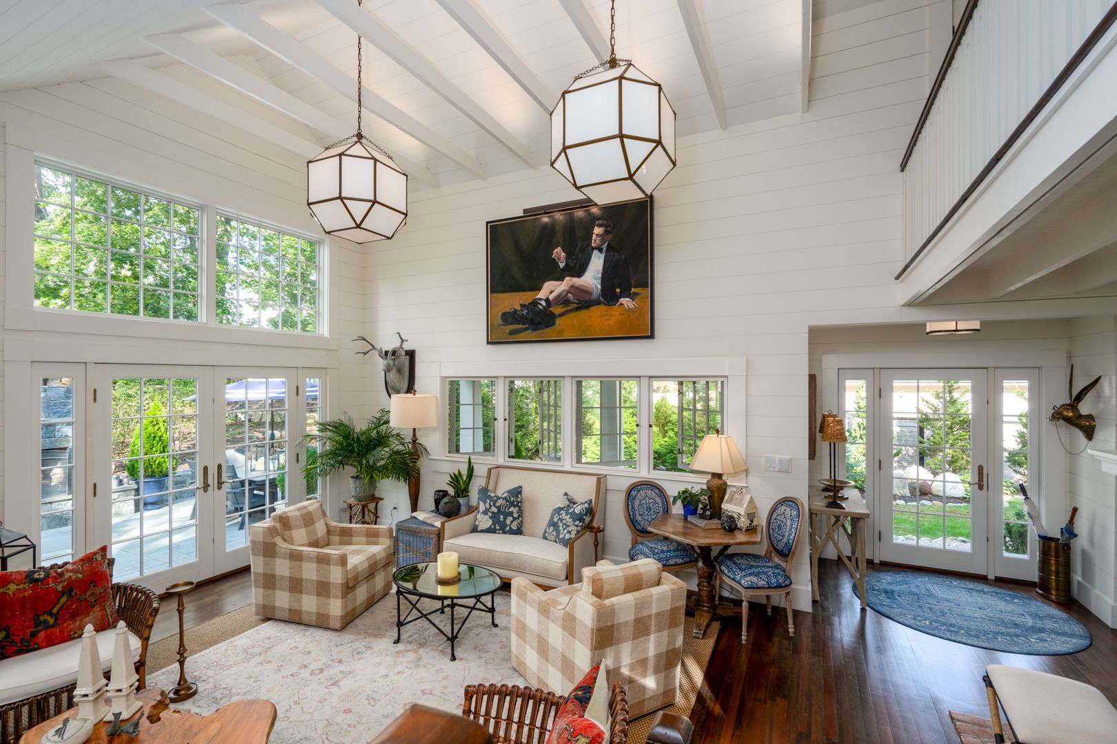 Bright sunroom with plaid armchairs, white beams, hanging lanterns, and large windows overlooking greenery