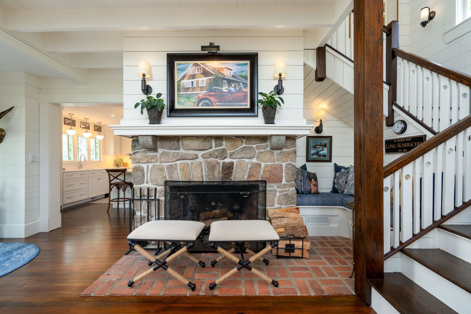Living room with stone fireplace, framed art, upholstered stools, and staircase with white railing
