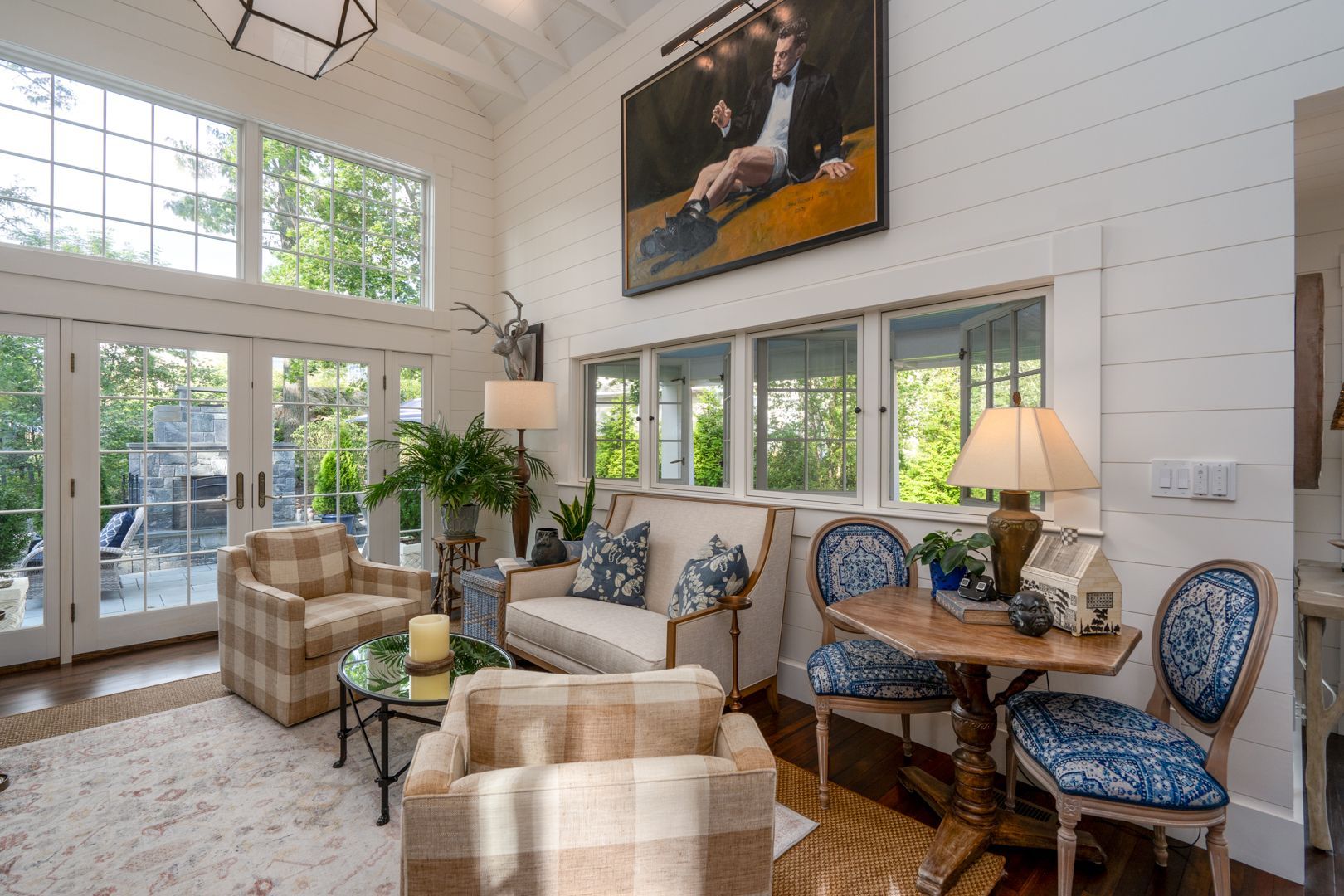 Bright sunroom with plaid chairs, blue-patterned seating, wood table, and large windows overlooking greenery