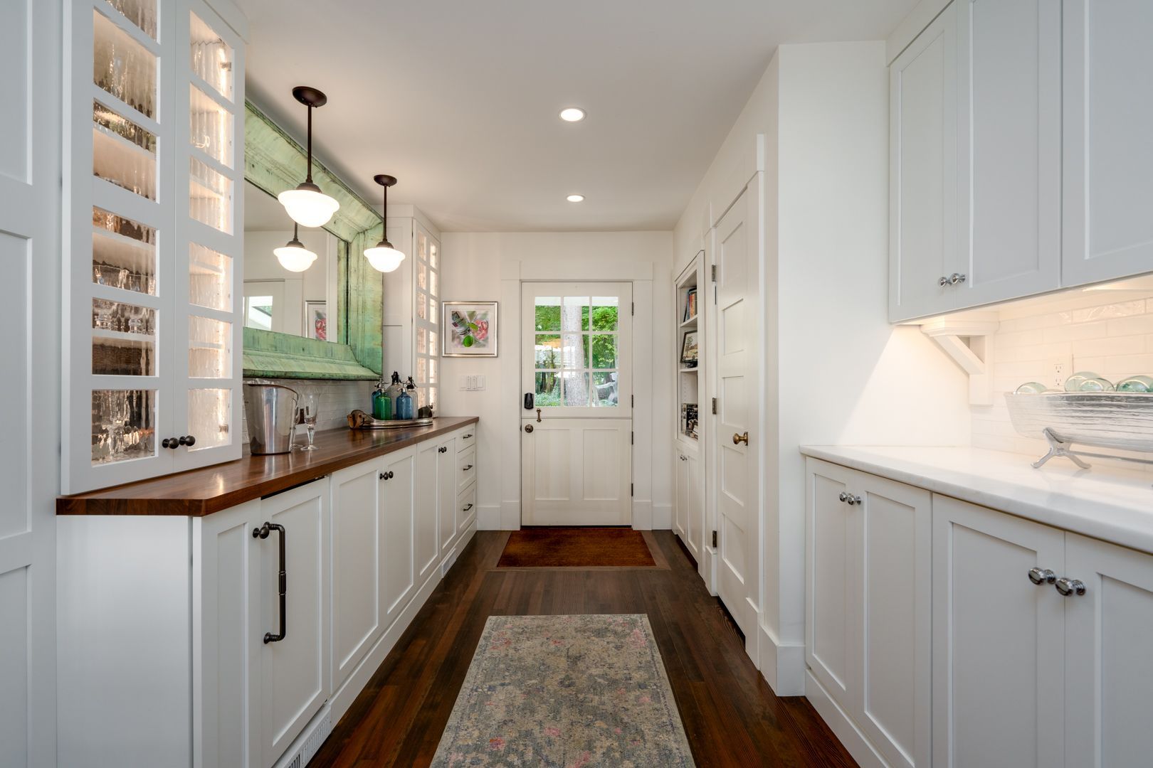 Bright galley kitchen with white cabinets, dark wood floor, and a rug leading to a back door.