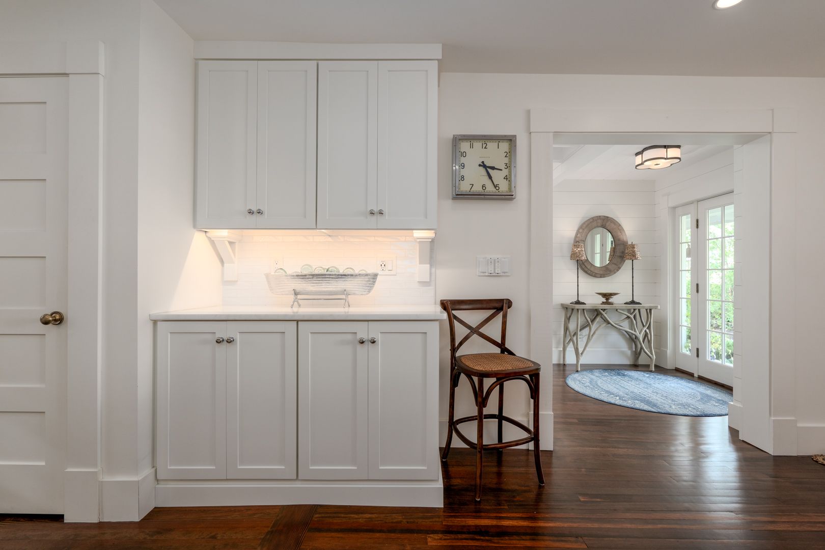 White hallway with built-in cabinets, wall clock, chair, and doorway to a bright room with rug.