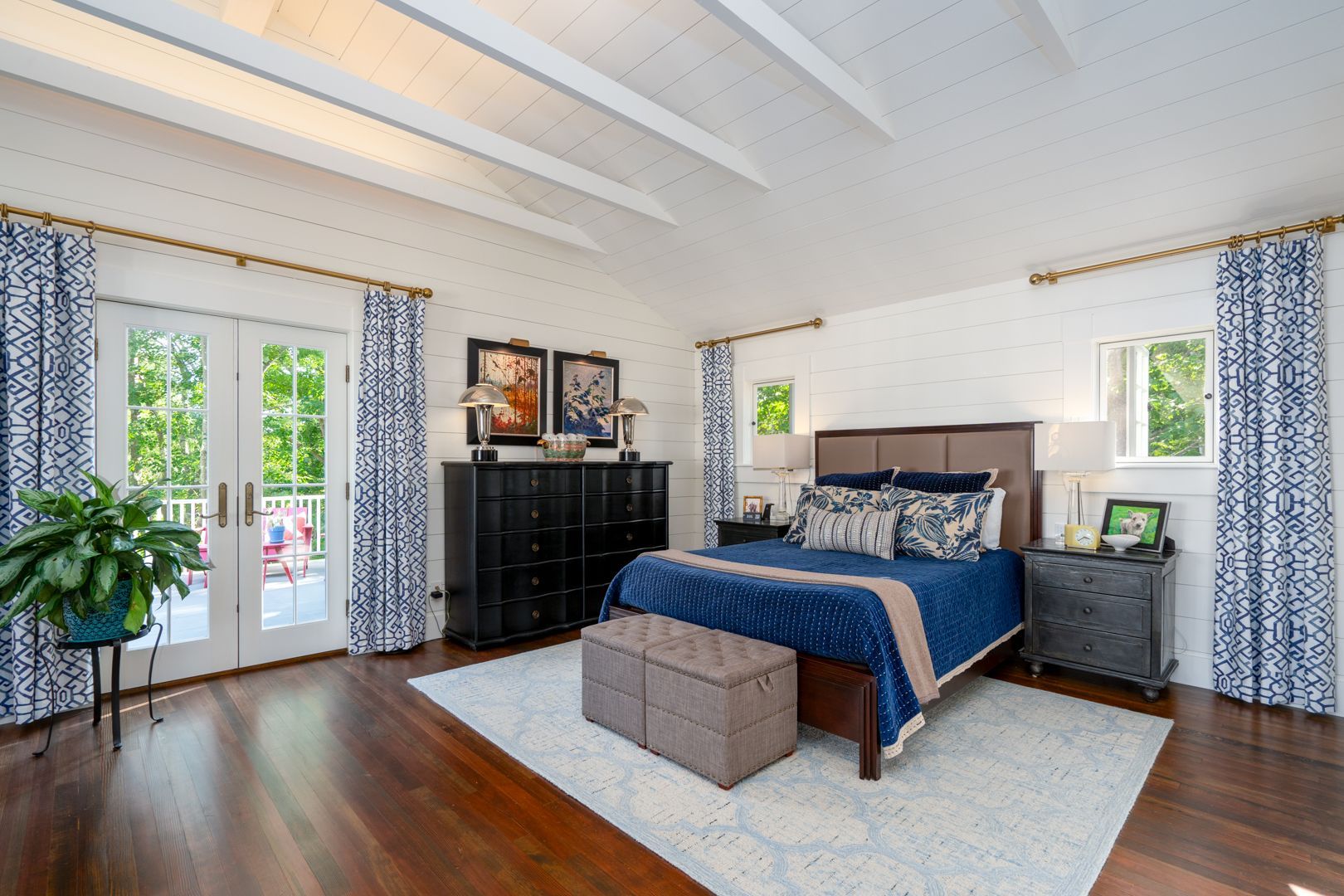 Bright bedroom with blue bedding, white ceiling beams, wood floors, and large windows with patterned curtains