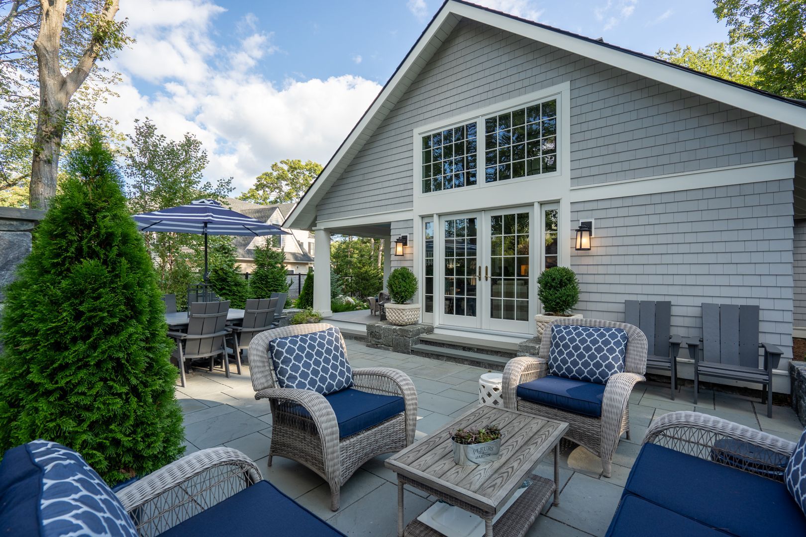 Patio seating area beside a gray house with wicker chairs, table, and umbrella under a blue sky