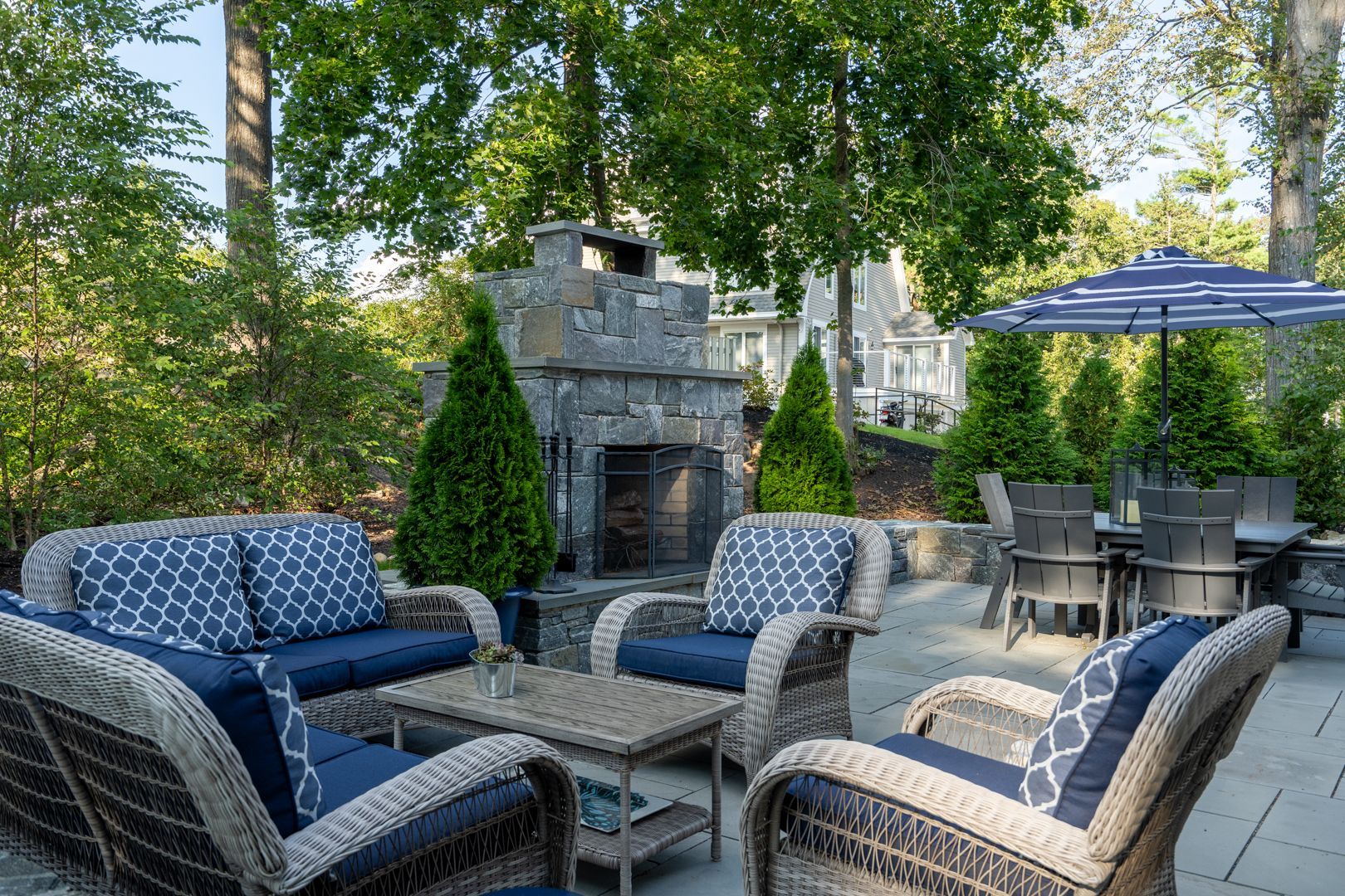Patio seating area with wicker chairs, stone fireplace, and blue umbrella under trees