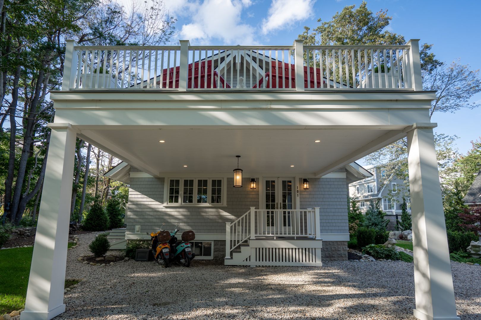 Front view of a white house with a raised porch, balcony railing, and gravel driveway under trees