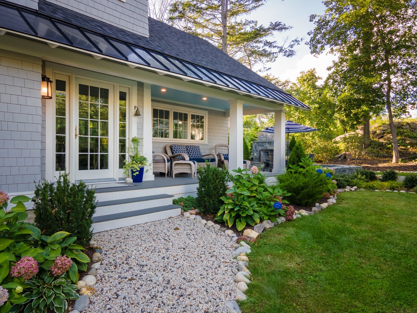 White porch with gray chairs, stone steps, and a gravel path beside a lush green lawn and garden.