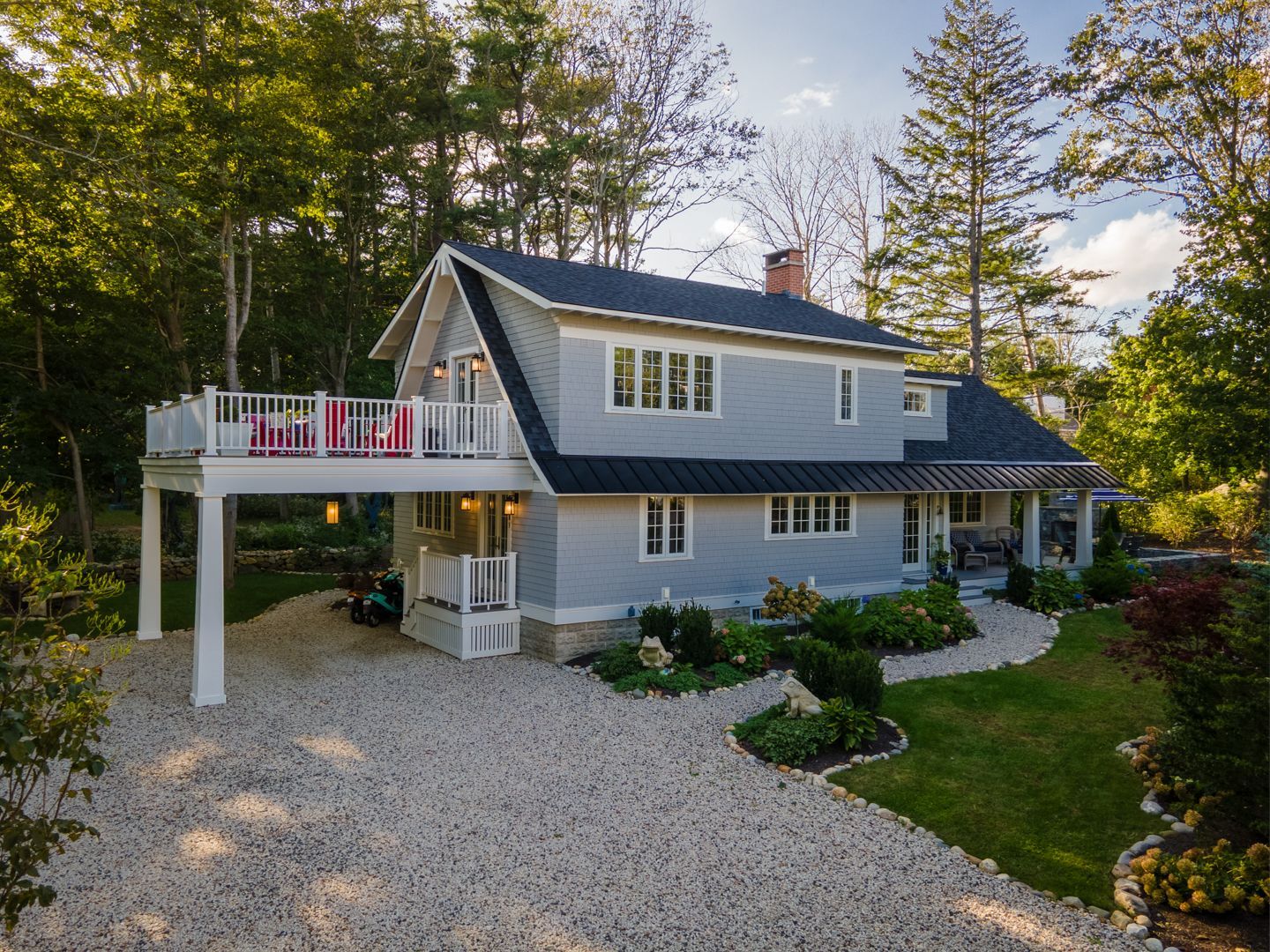 Gray two-story house with white trim, deck, and gravel driveway surrounded by trees and garden landscaping