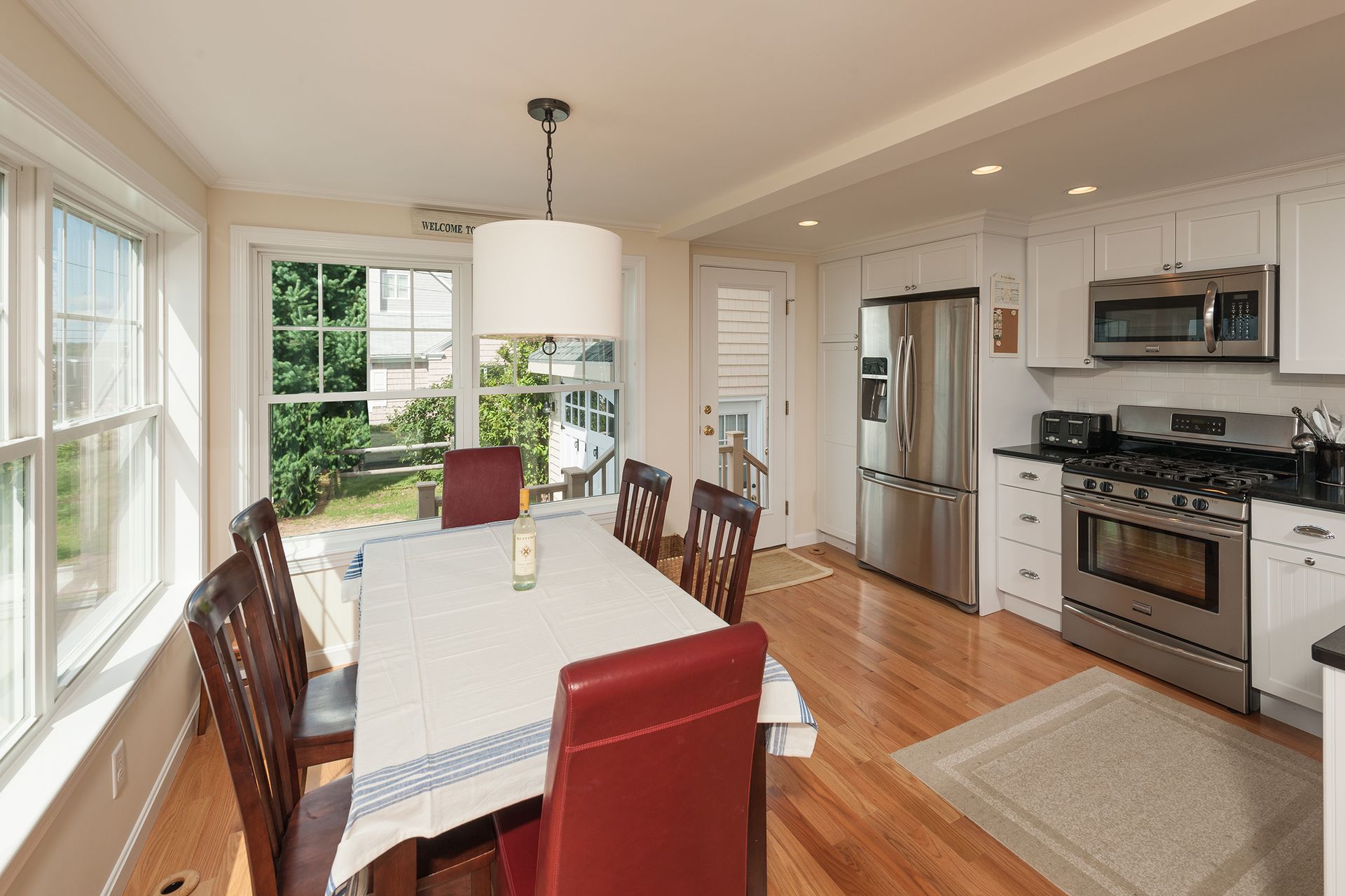 Bright dining area and kitchen with white table, red chairs, hardwood floors, and large windows.