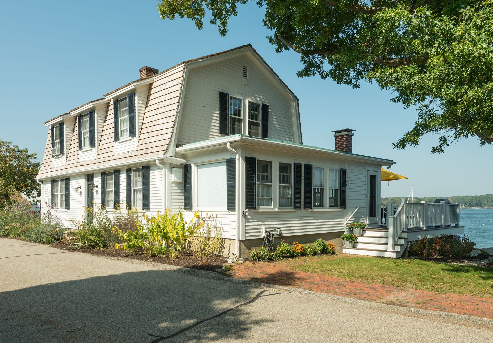 White farmhouse-style house with porch and green shutters beside the water on a sunny day