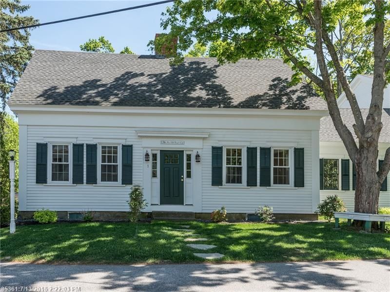 White house with gray roof, black shutters, and a front door under large trees in bright daylight