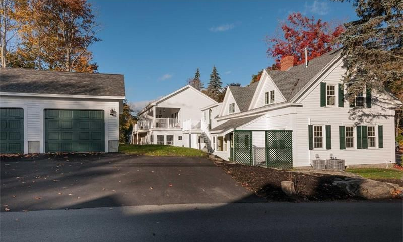 White houses and a garage along a paved driveway in a quiet residential neighborhood, with autumn trees.