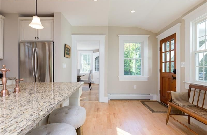 Bright kitchen with granite island, stainless fridge, wood floors, and glass front door to a hall.