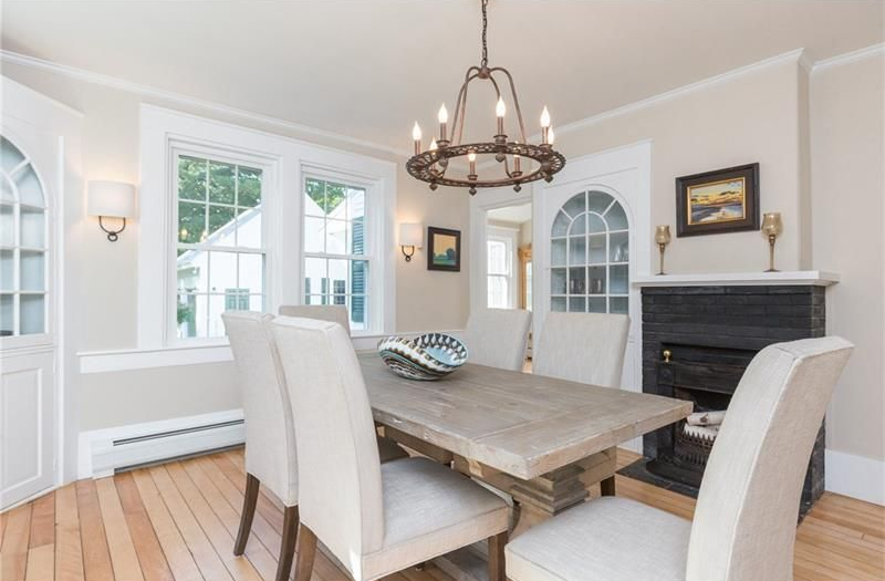 Bright dining room with white chairs, rustic wood table, chandelier, and fireplace near large windows