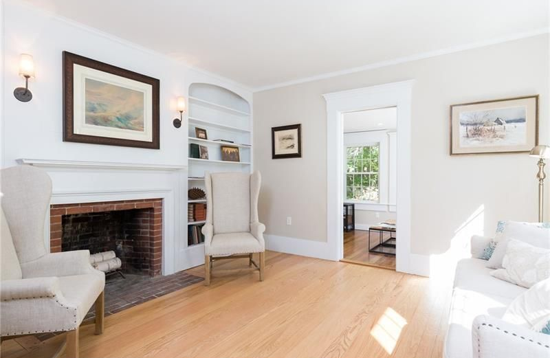Bright living room with white furniture, a brick fireplace, hardwood floors, and an open doorway to another room