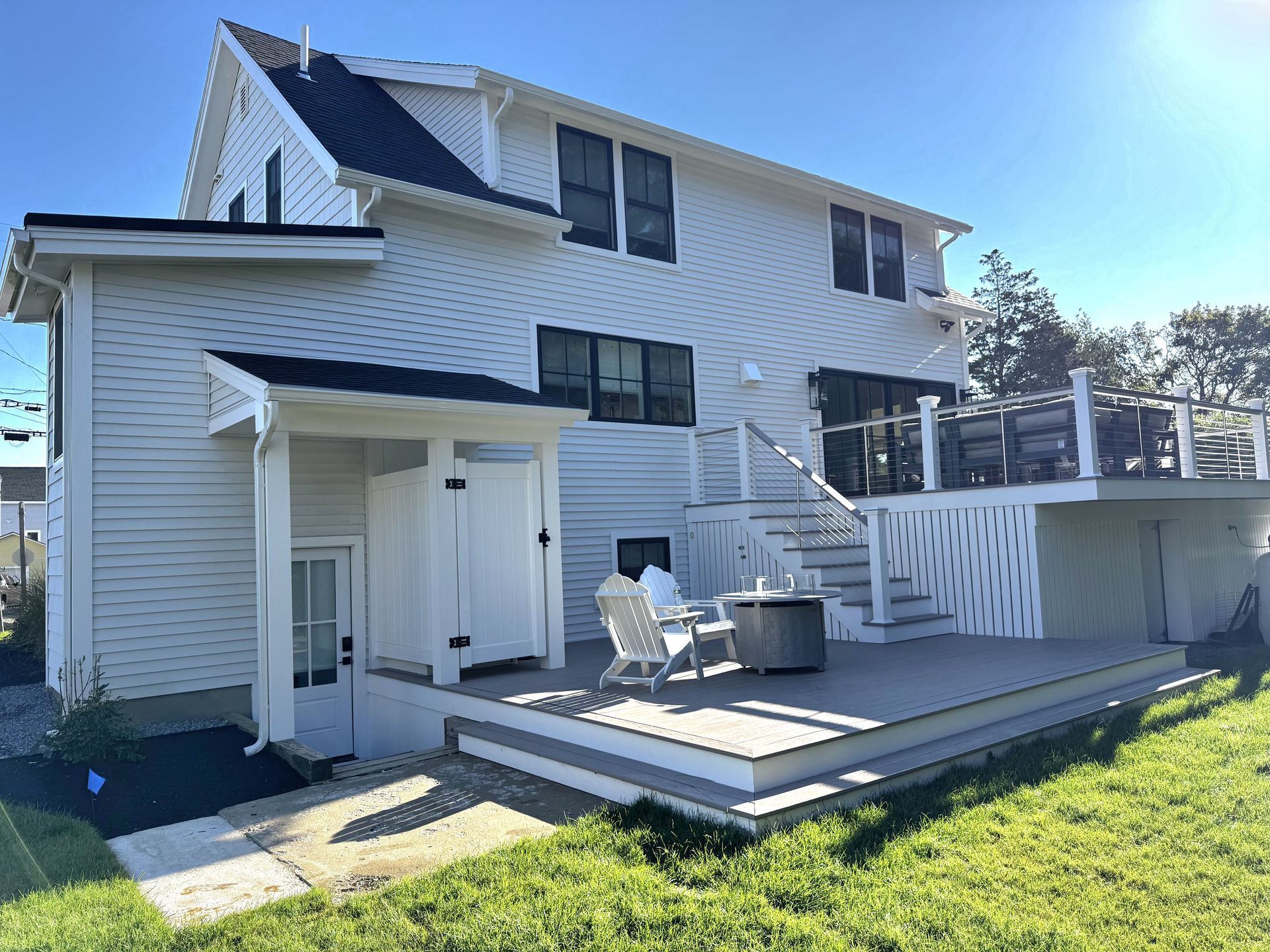 White two-story house with a small deck, stairs, patio furniture, and a grassy yard in sunlight