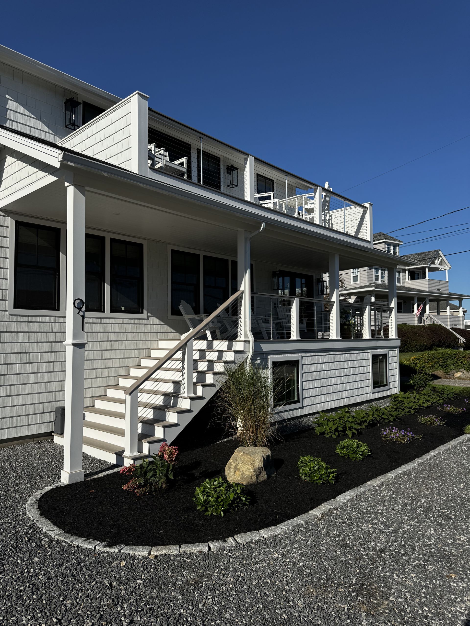 White coastal house with gray siding, front stairs, and a landscaped gravel yard under a clear blue sky