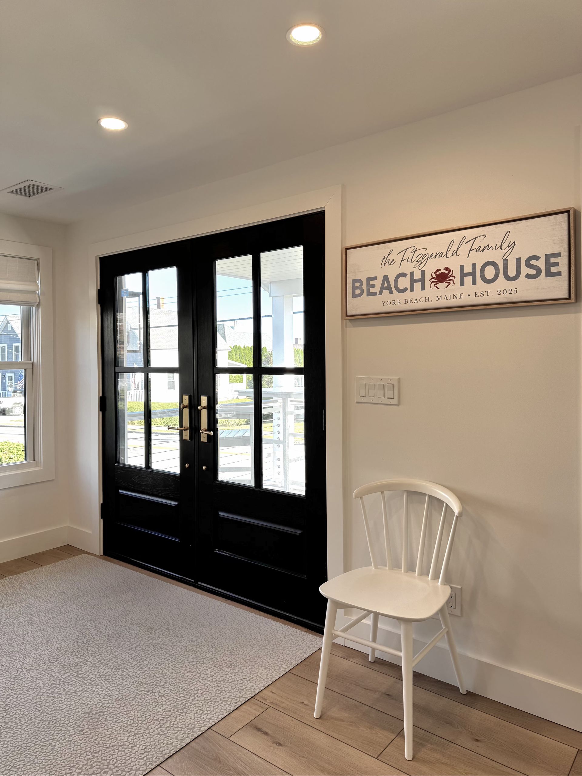 Bright entryway with black French doors, white chair, and a Beach House sign on the wall.