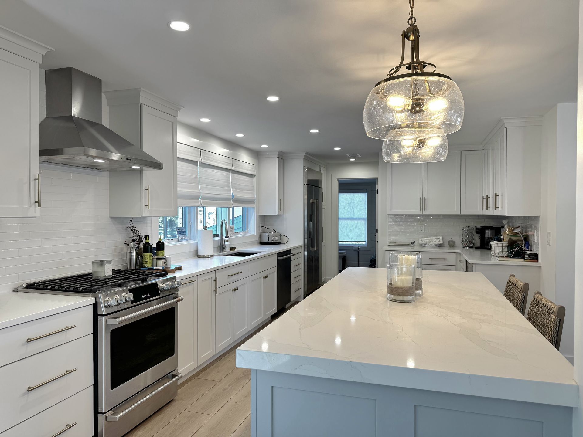 Bright modern white kitchen with island, pendant light, stainless appliances, and large windows