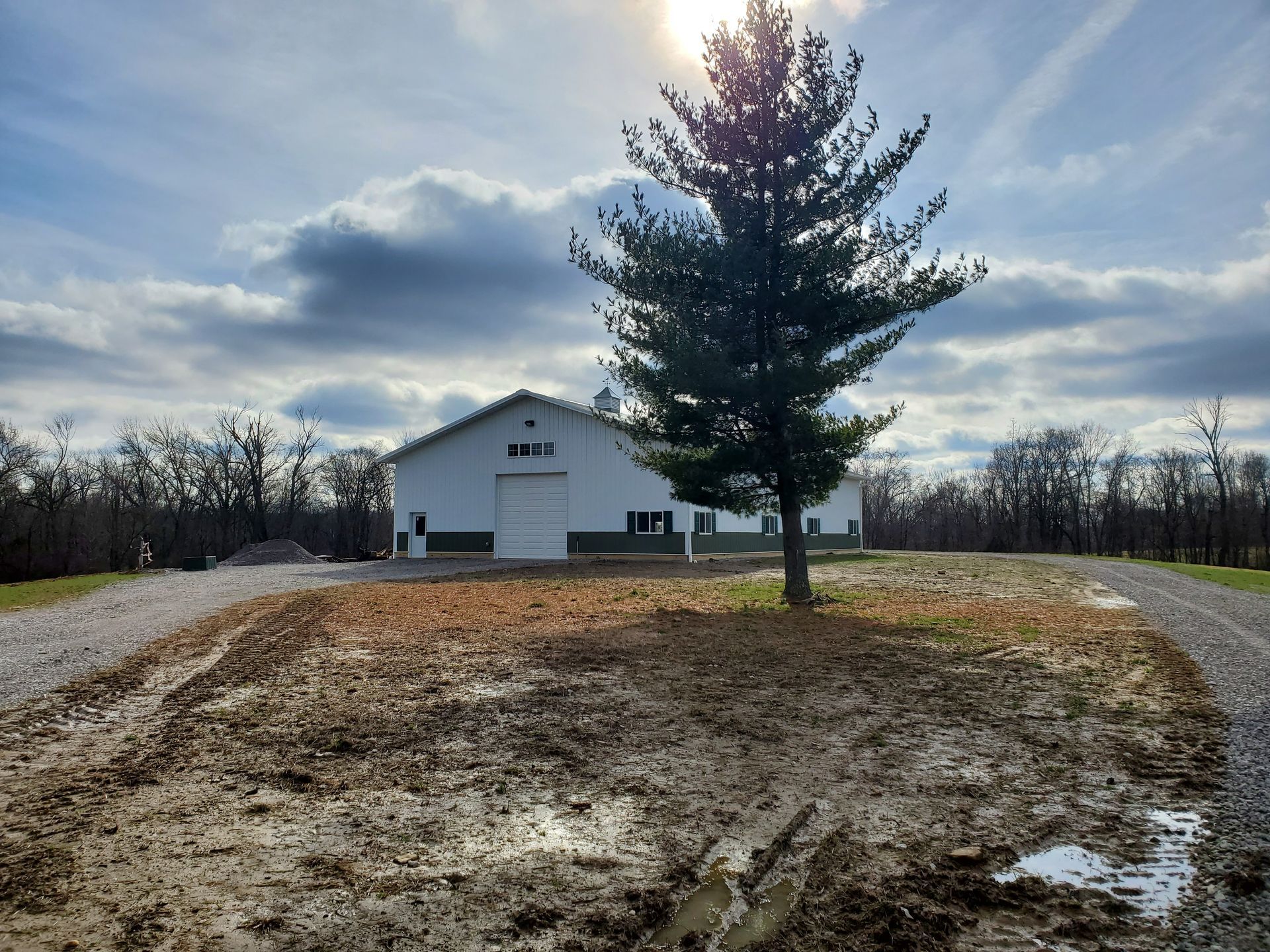Barn with a tree in front, cloudy sky, muddy ground.