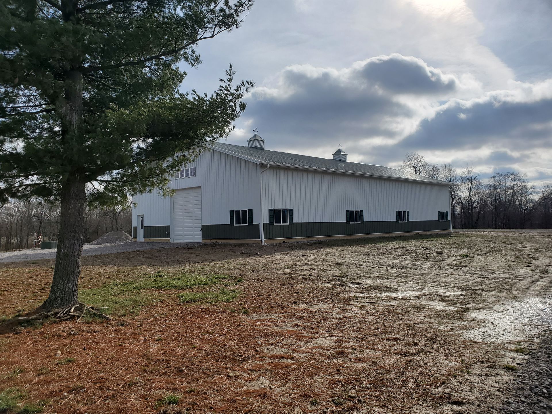 White barn with green trim and a closed door on a cloudy day. A tree stands to the left.