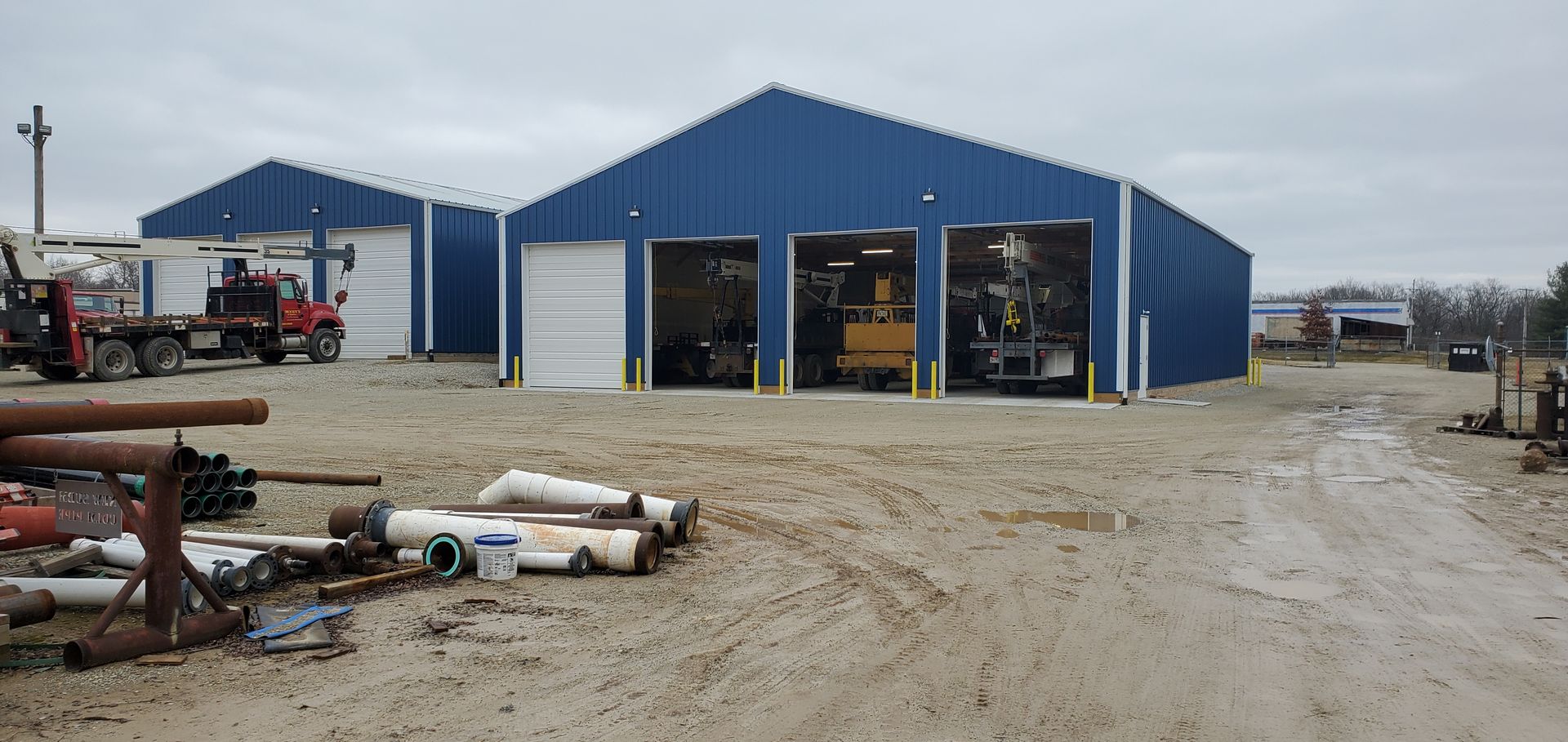 Blue metal buildings with garage doors, vehicles, and equipment on gravel lot.