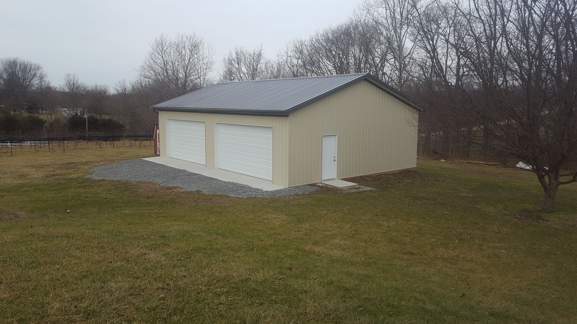 Tan metal garage with two bays, a small door, and a gravel base on a grassy lot.