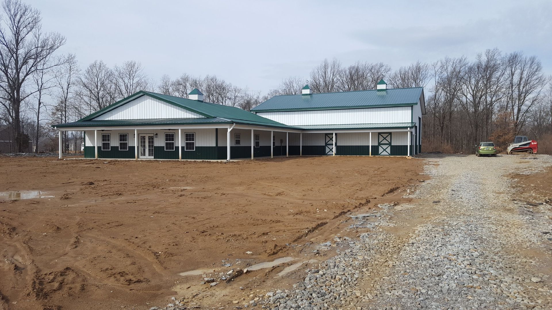 A white and green horse barn on a dirt lot with a gravel driveway.