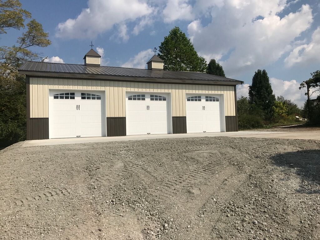 Three-bay garage with white doors, tan siding, and a gravel driveway against a blue sky with clouds.