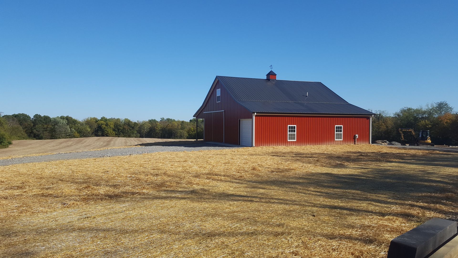Red barn with a dark roof on a sunny day, set in a field with trees in the background.