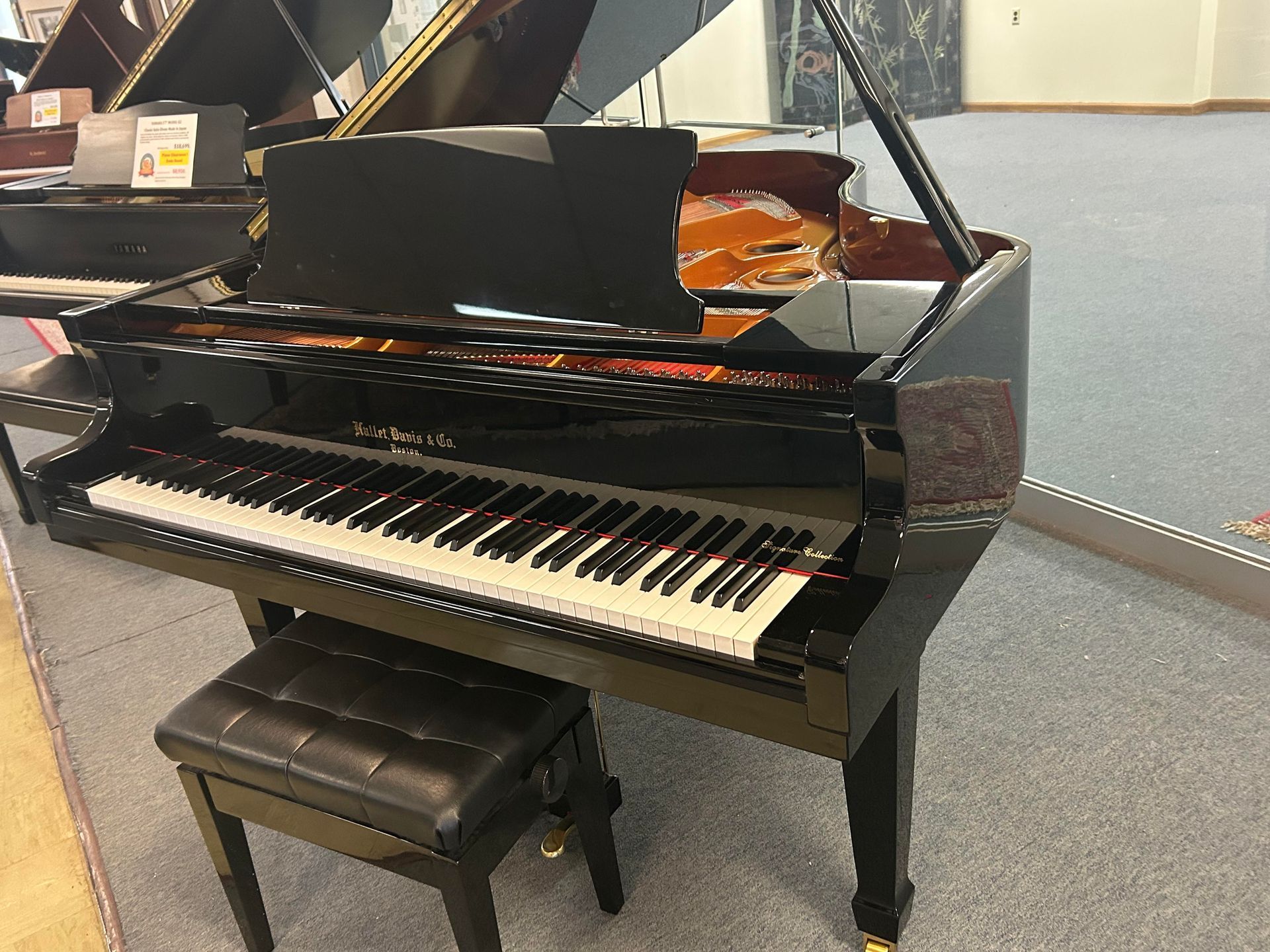 A black grand piano with its lid open, sitting on a grey carpeted floor with a matching padded bench.