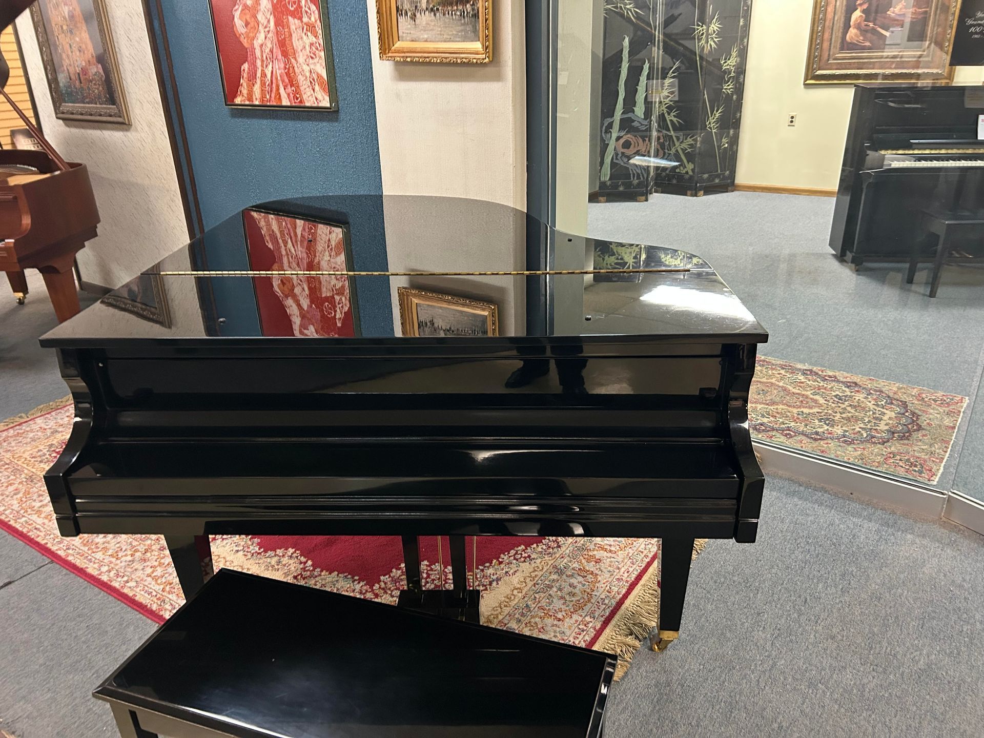 Black grand piano in a showroom, with matching bench. Reflective surface. Red and patterned rug.